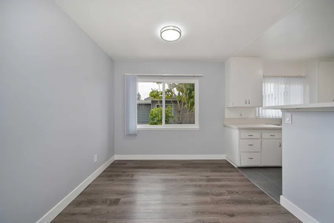 A brightly lit room with light gray walls and modern flooring. A window with white curtains offers views of greenery outside. The space features a small kitchen area with white cabinetry, creating a clean and open atmosphere, ideal for living or dining.