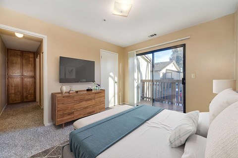 A bedroom with eggshell-colored walls, carpet, and a sliding glass door to a balcony. The room is staged with a bed, a dresser, and a TV.