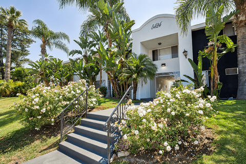 Entrance of the community with stairs leading up to the white building. The complex is surrounded with grass, trees, and flowers.