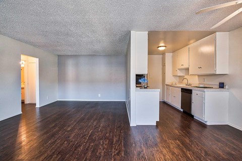 Overview of the living room and kitchen. Floors are wood style vinyl and the walls are white.
