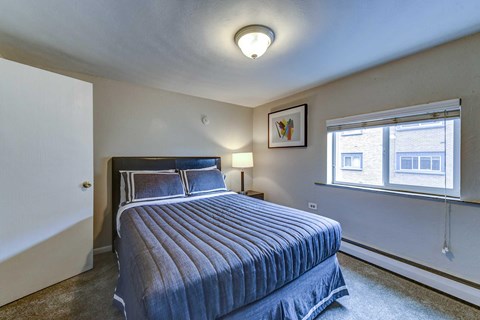 A bedroom with a bed, a nightstand with a lamp, and a window.