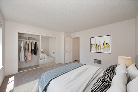 A bedroom with carpet, white walls, and a sliding mirrored closet door. The room is staged with clothes in the closet, a bed, and a rug.