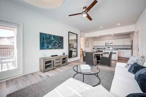 A modern living room with wood style plank flooring, a ceiling fan, and white walls. The room is staged with a dining room table, a couch, a rug, and a tv.