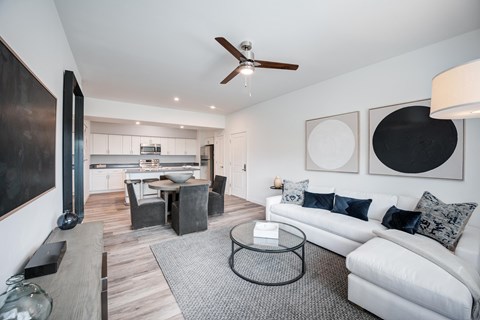 A modern living room with wood style plank flooring, a ceiling fan, and white walls. The room is staged with a dining room table, a couch, a rug, and a tv.