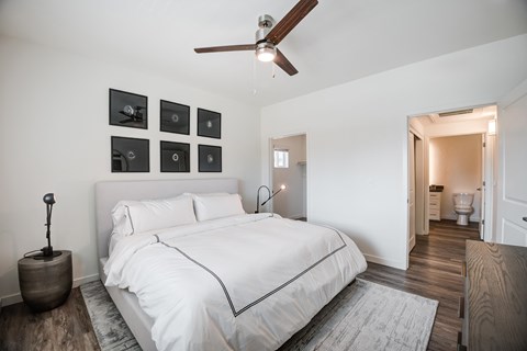 A modern bedroom with wood style plank flooring, white walls. and a ceiling fan. The room is staged with a rug, bed, and a nightstand.