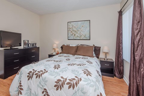 A bedroom premium wood style flooring, beige walls, and a window. The room is staged with a bed, nightstands, a television, and a painting on the wall.