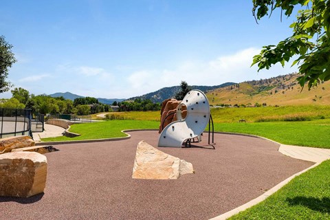 A playground with a white curved climbing wall and a brown rock in a green space.