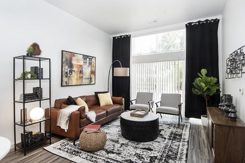 a living room with a couch, coffee table, two chairs, and lamp and plant in front of large windows with drapes