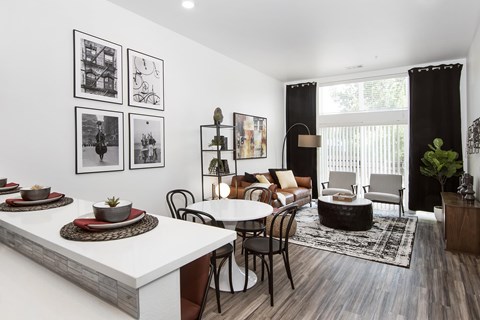a view from the kitchen looking past a counter into the living room that is furnished with a table and couch in front of a window