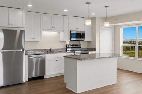 A modern kitchen with white cabinets, stainless steel appliances, and stone countertops with a view of the mountains through oversized windows at The Beaumont Apartments, Bozeman