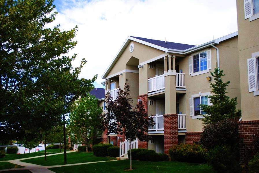 Country Oaks Apartment building with half-wall red brick work, white balconies, and tree lined pathways on a sunny day with a blue sky and white clouds at Country Oaks Apartments in Clearfield, 84015