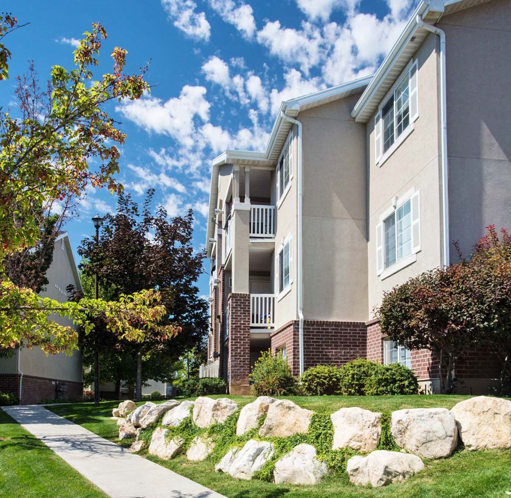 Side view of Country Oaks Apartment building with columns featuring part brick, large windows, white balcony railings, and pathway with rock landscaped yard area at Country Oaks in Clearfield, UT