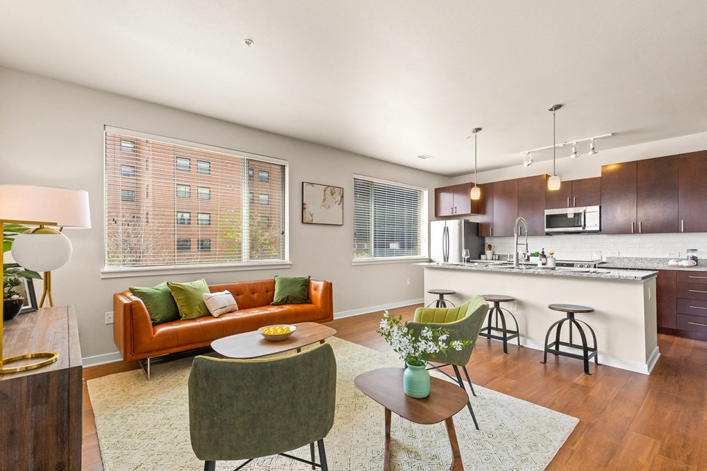 Modern living room with an orange couch, green accent chairs and oversized windows, complete with view into kitchen with barstool seating and pendant lighting at Sylvan Uptown, Denver, CO, 80203