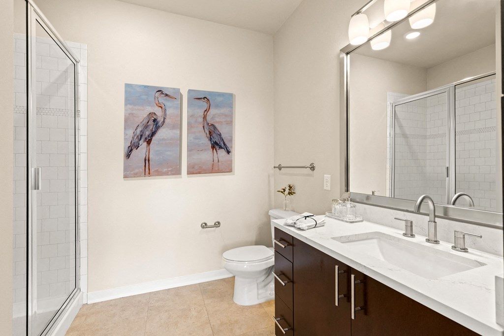View into the bathrom with a glass enclosed shower and well-lit vanity area with a large mirror at Sylvan Uptown, Denver, CO