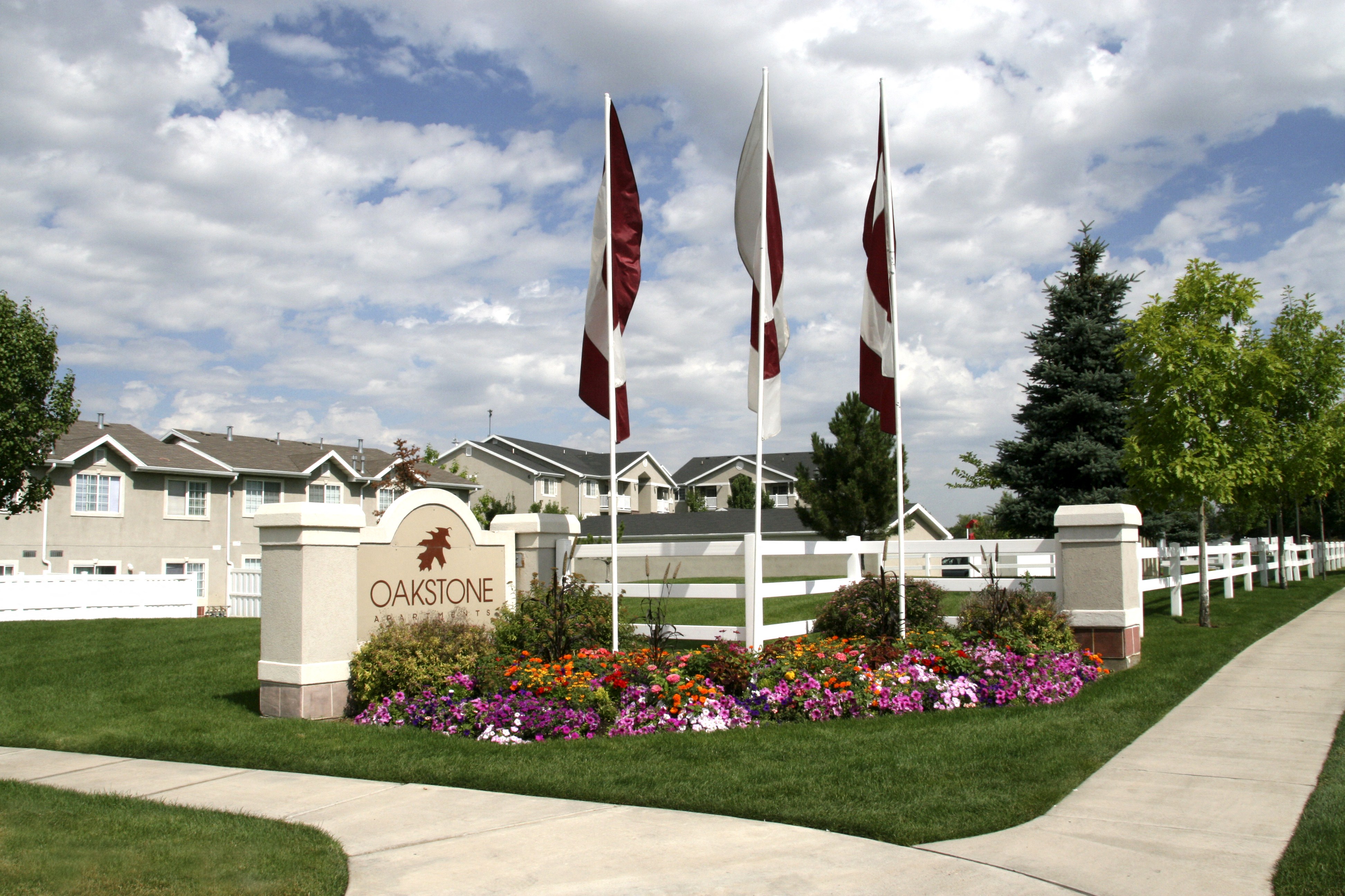View of the front of Oakstone Apartments with property signage, three flags, and manicured brightly-colored flowers and walking pathways through the green grass at Oakstone Apartments in Utah 84015