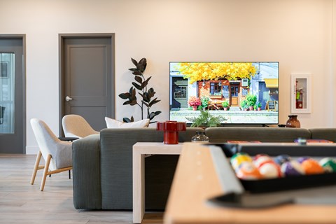 A living room with a grey sofa, a white chair, a wooden table, and a television displaying a yellow building.