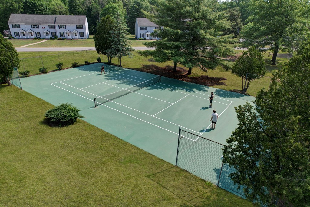 Two people playing tennis on a court with trees and houses in the background.