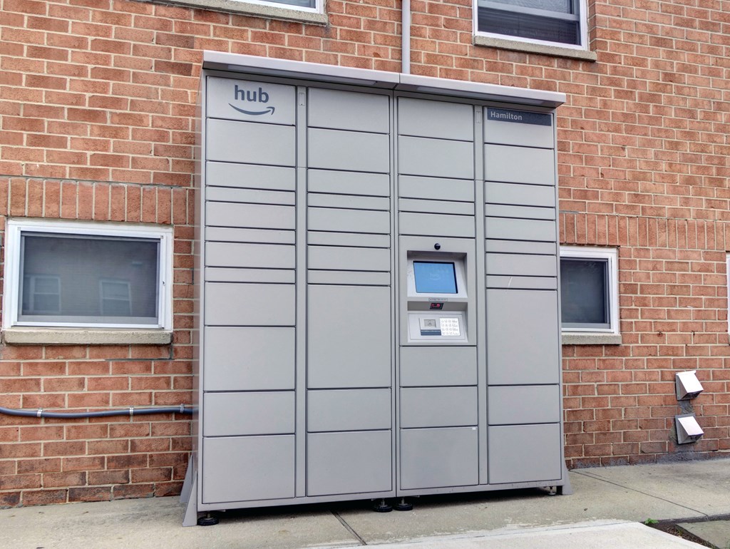 A grey Amazon Locker is on the sidewalk in front of a brick building.