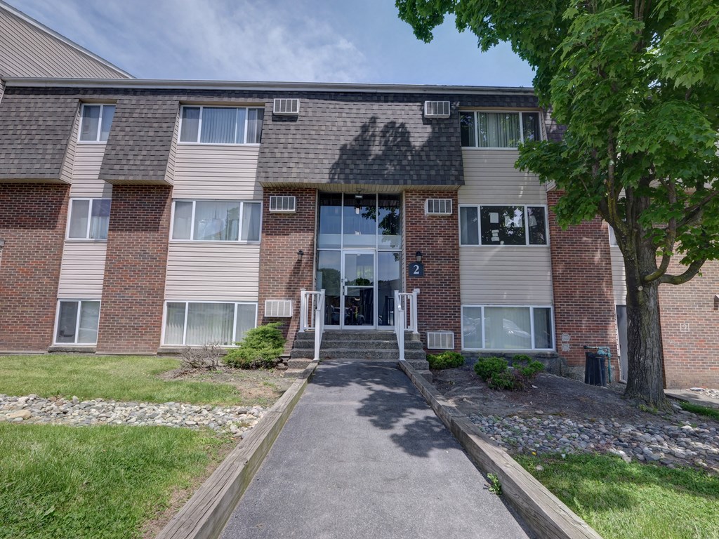front view of a brick apartment building with a walkway and grass at Highland Club apartments, Watervliet, NY 12189