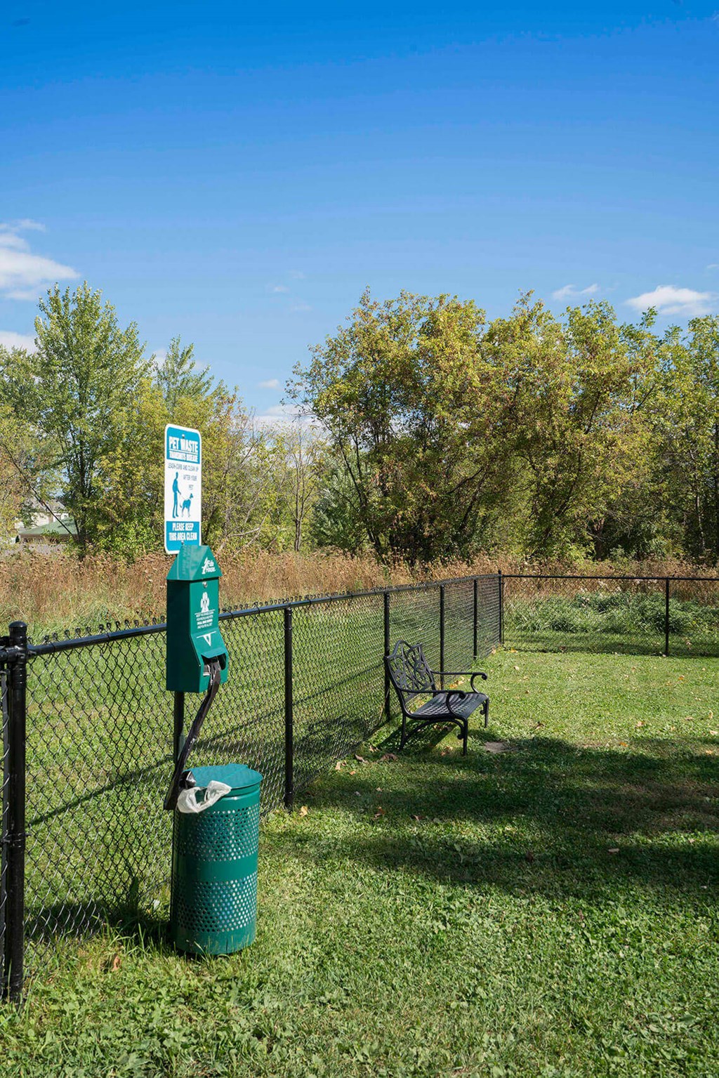 a park bench and trash can in front of a fence with trees in the background
