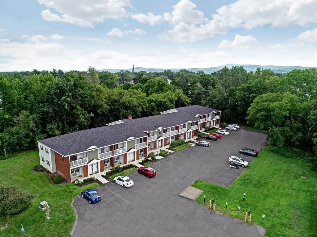 an aerial view of a red brick building with a gray roof and a parking lot in front