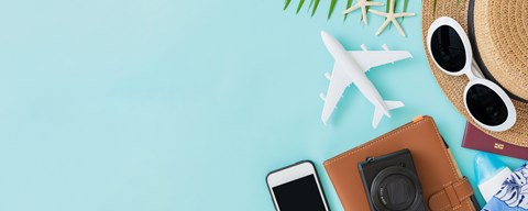 A flat lay of travel items including a hat, sunglasses, a camera, a passport, a phone, and a small airplane model on a blue background.