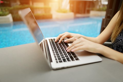 A person is typing on a laptop by a poolside.