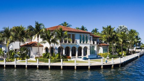 A large white house with a red roof is situated on a dock with palm trees in the background.