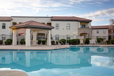 A swimming pool in front of a building with a patio and chairs.