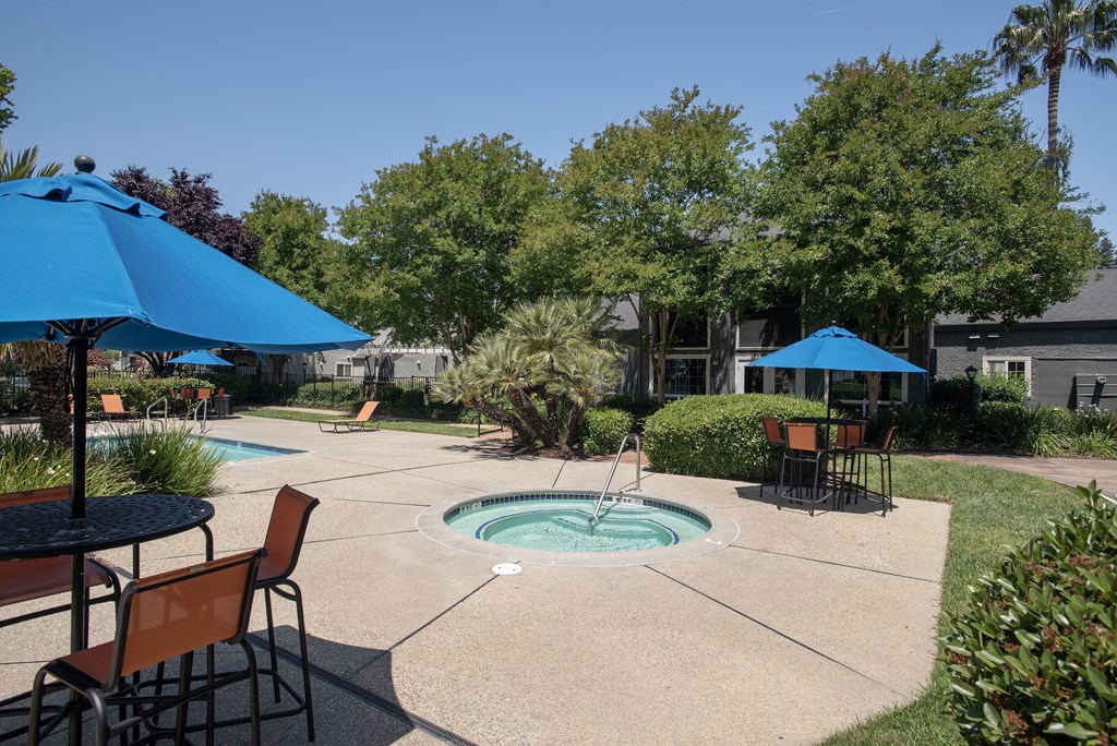 outdoor spa with manicured landscaping and patio tables with orange chairs and blue umbrellas at Woodbridge Apartments, California