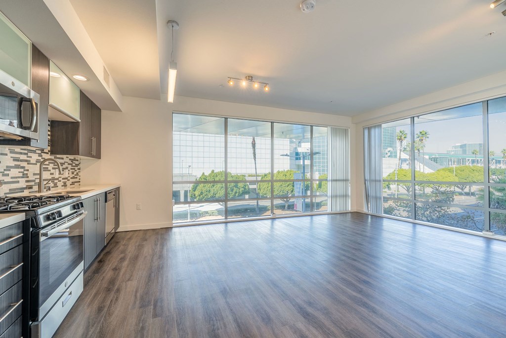 an empty living room with a large window and a kitchen with stainless steel appliances