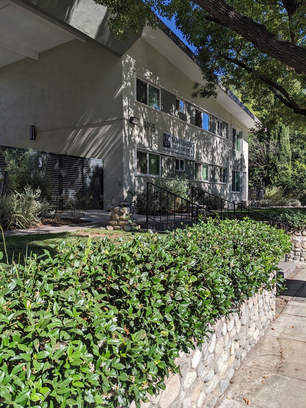 Sidewalk view of stone wall garden beds and mature shade trees in front of Los Robles Apartments in Pasadena, California.