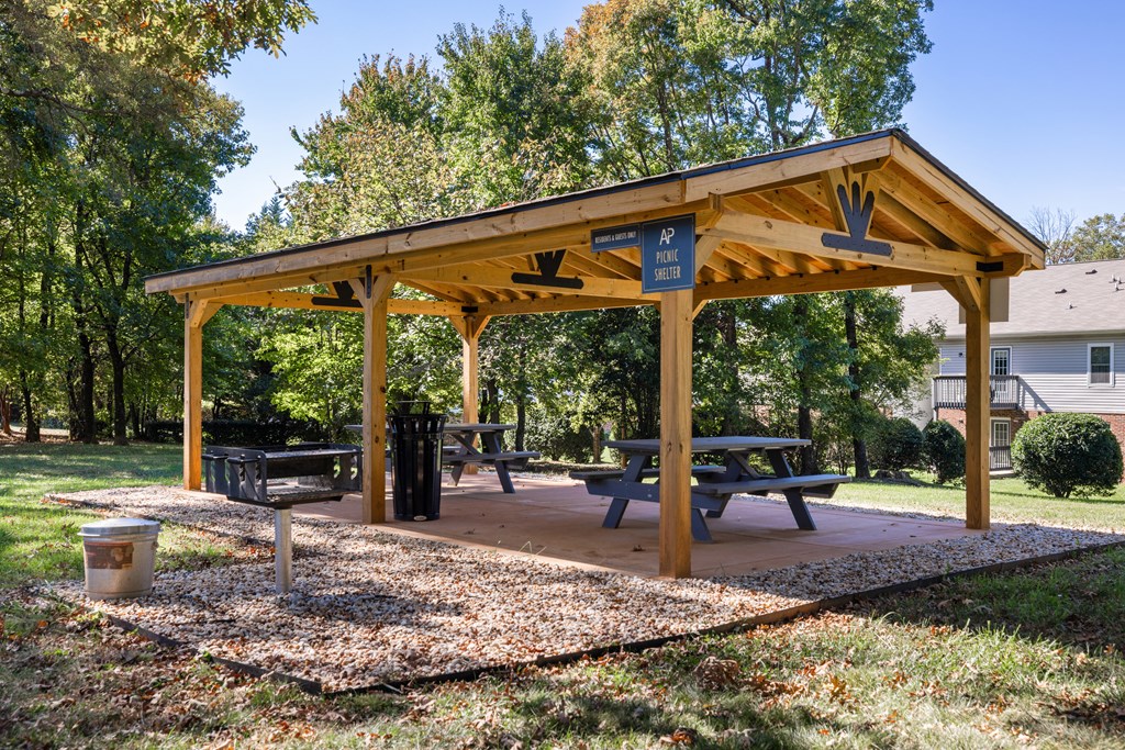 A wooden pavilion with a picnic table and benches is surrounded by trees.