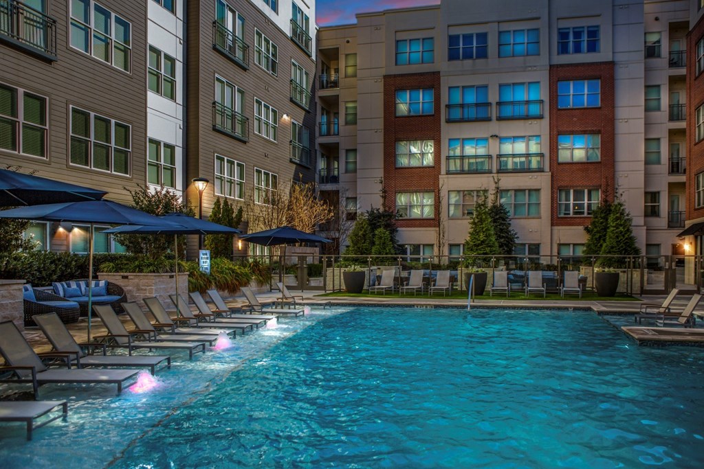 A swimming pool surrounded by lounge chairs and umbrellas in front of apartment buildings.