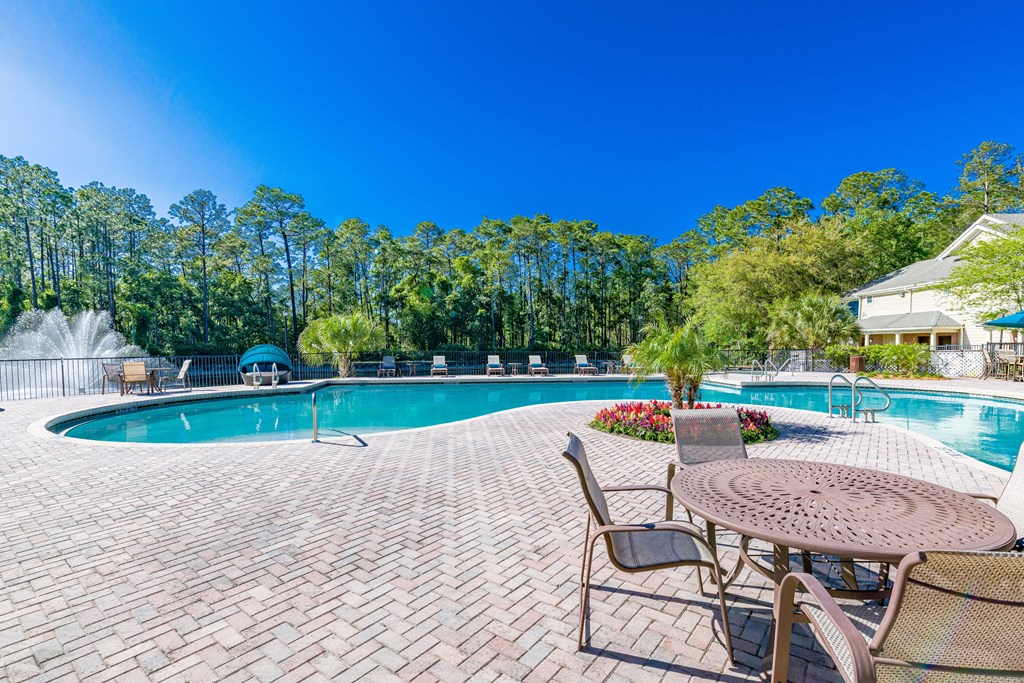 A table with chairs around it and a pool with trees in the background.