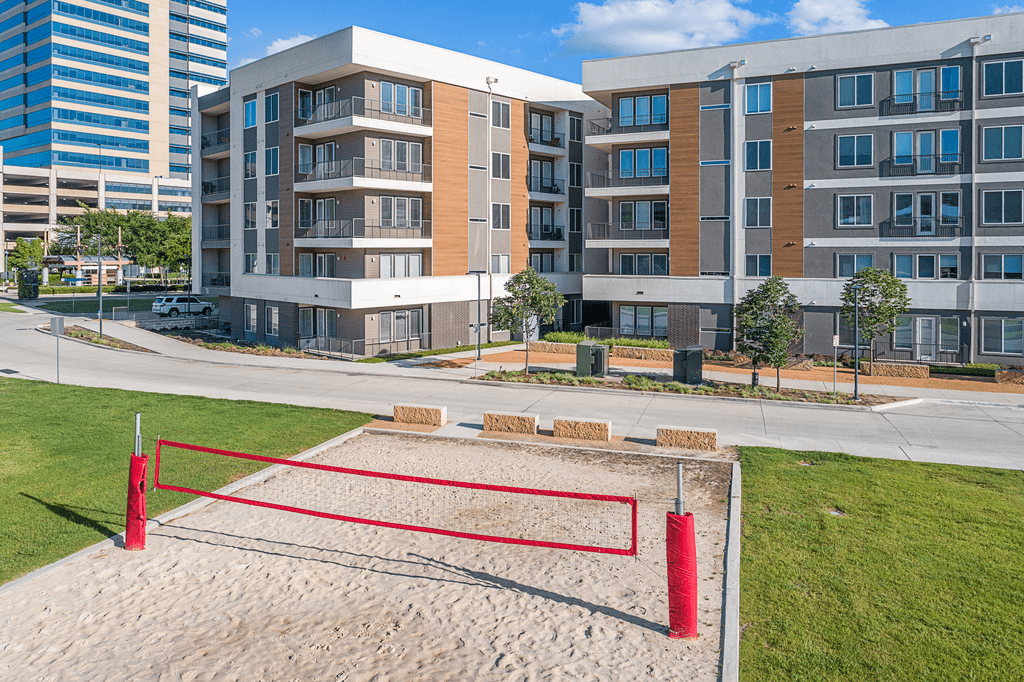 A red volleyball net is set up in the sand in front of apartment buildings.
