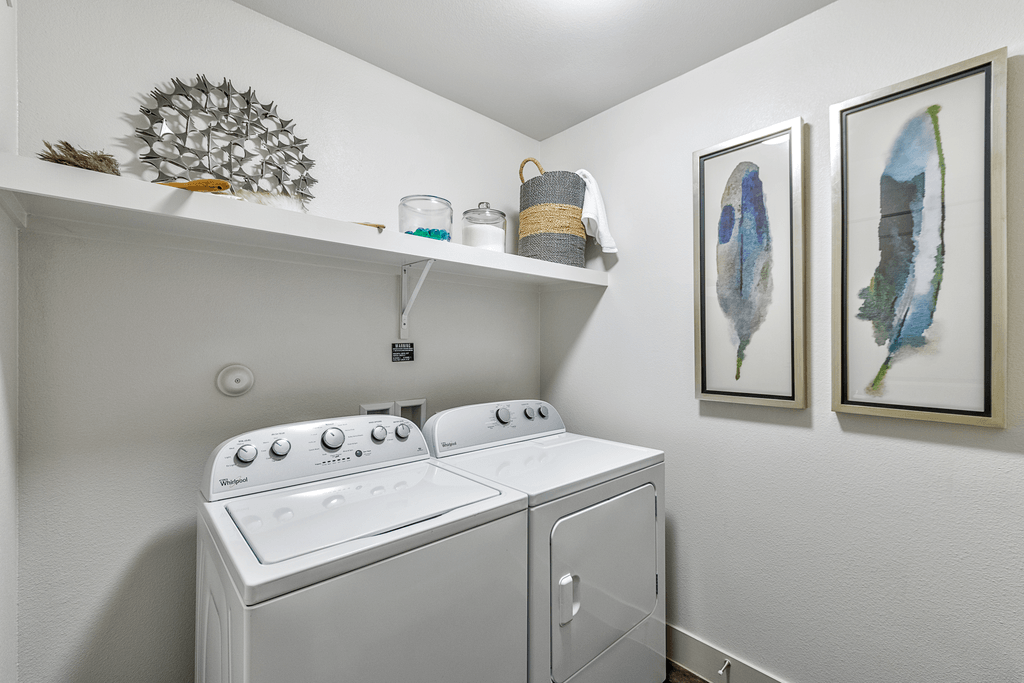 A white washing machine sits in a laundry room with two framed pictures on the wall.