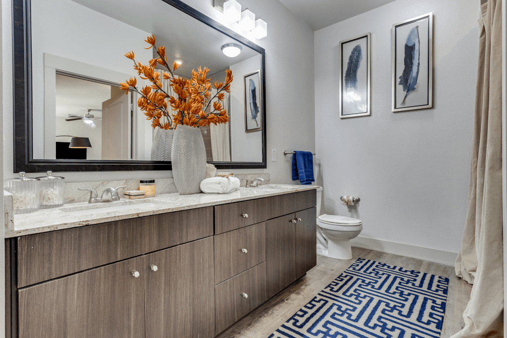 A bathroom with a large mirror, a vase with orange flowers, and a blue and white patterned rug.