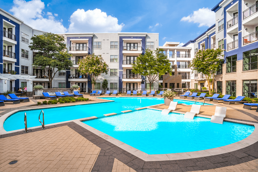 A large swimming pool surrounded by lounge chairs and trees in front of apartment buildings.