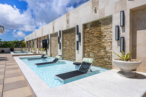 A pool area with a stone wall and lounge chairs.