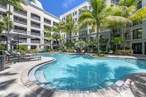 A large outdoor swimming pool surrounded by palm trees and apartment buildings.