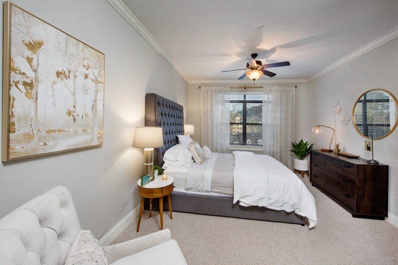 A bedroom with a bed, a chair, a side table, a ceiling fan, and a painting on the wall.