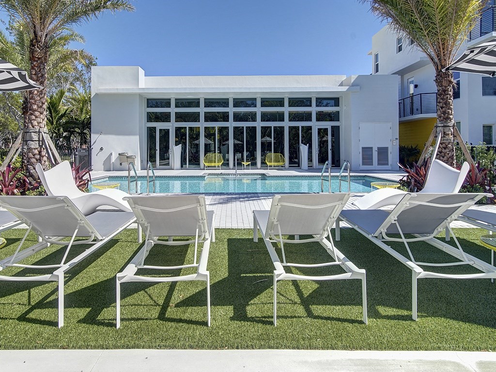 A row of white chairs are set up on a lawn in front of a pool and a white building.