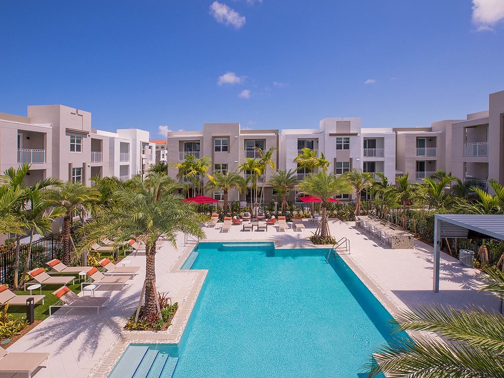 A swimming pool surrounded by palm trees and sun loungers in front of apartment buildings.