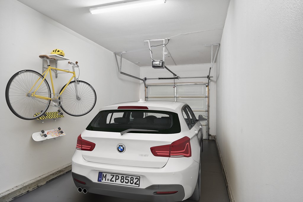 A white BMW car is parked in a narrow garage with a yellow bicycle hanging on the wall above it.