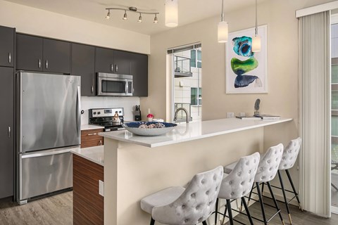 A modern kitchen with a refrigerator, microwave, and a counter with four white chairs.