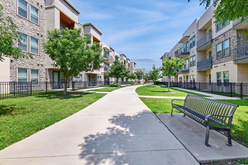 A long concrete walkway with a bench on the right side and a row of apartment buildings on the left.