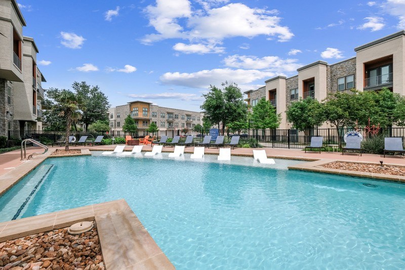 A swimming pool surrounded by lounge chairs and trees in front of apartment buildings.