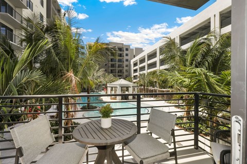 A table with chairs is on a balcony with a pool and palm trees in the background.