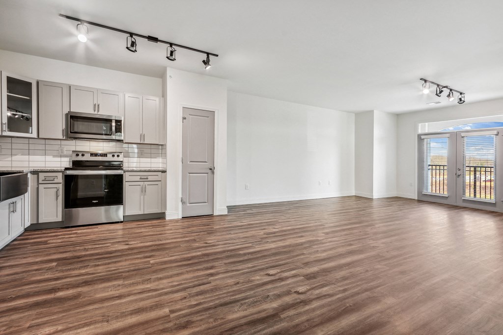 an empty living room with wood flooring and a kitchen with stainless steel appliances